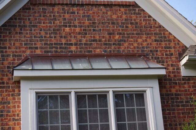 A close-up of a house exterior with red brick siding, featuring a copper awning above a set of windows.
