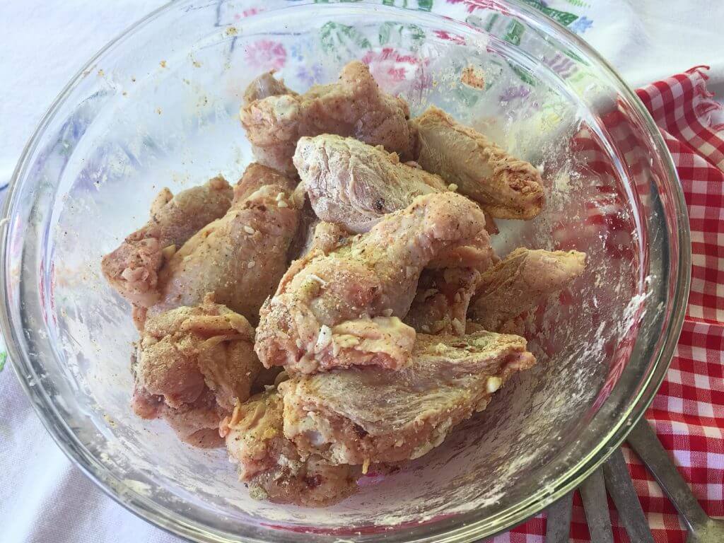 A glass bowl filled with seasoned raw chicken wings, ready for cooking, is placed on a table with a red and white checkered cloth nearby.