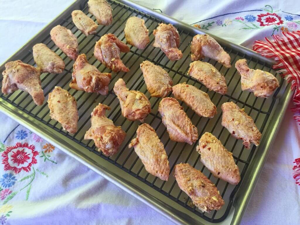 A baking tray with a wire rack holds rows of seasoned raw chicken wings, spaced evenly and ready for baking, with a floral-patterned tablecloth underneath.