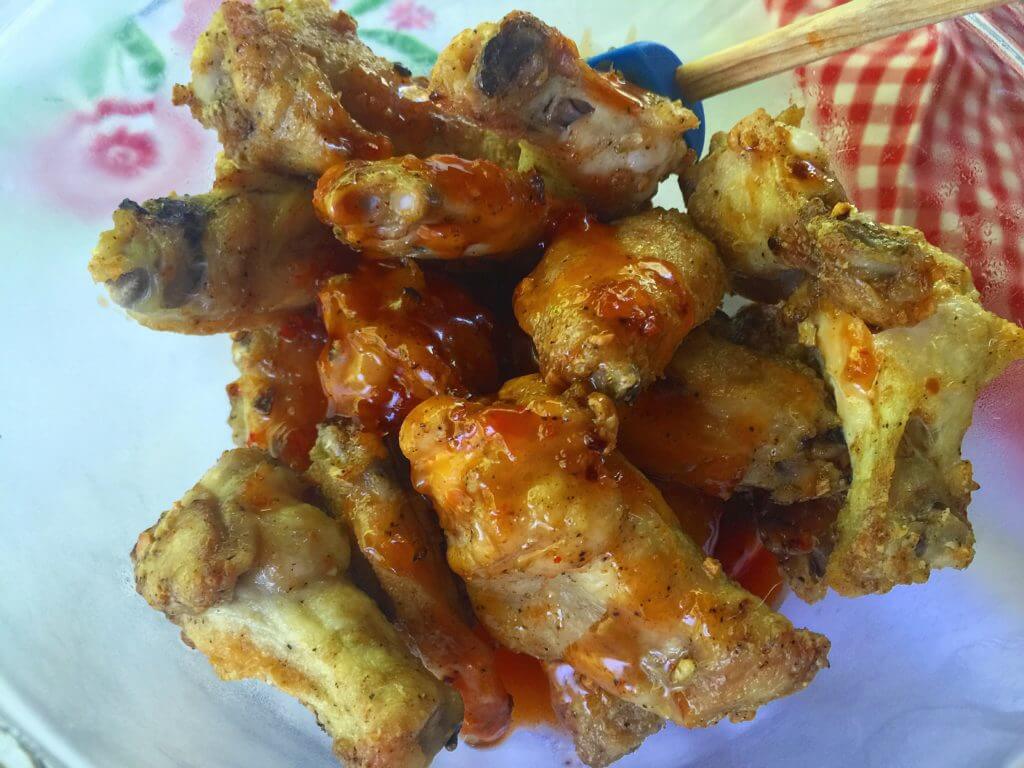 A bowl of cooked chicken wings coated in a glossy red sauce is set on a table with a floral tablecloth and a red and white checkered cloth in the background.