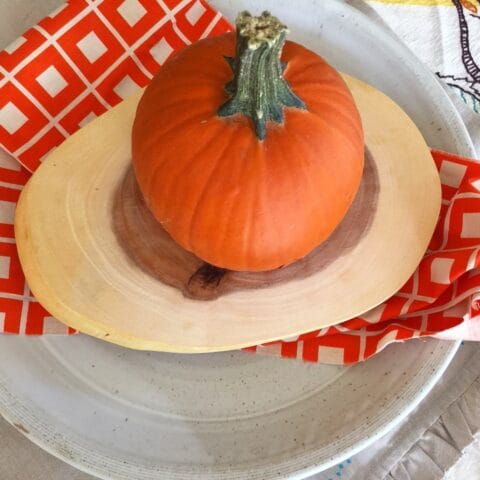 A small pumpkin sits on a wooden cutting board, placed on a plate with a red and white patterned napkin underneath.