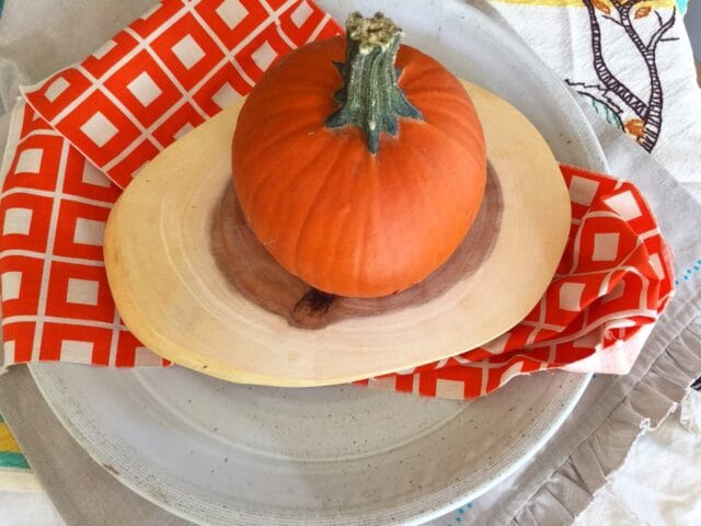 A small pumpkin sits on a wooden cutting board, placed on a plate with a red and white patterned napkin underneath.