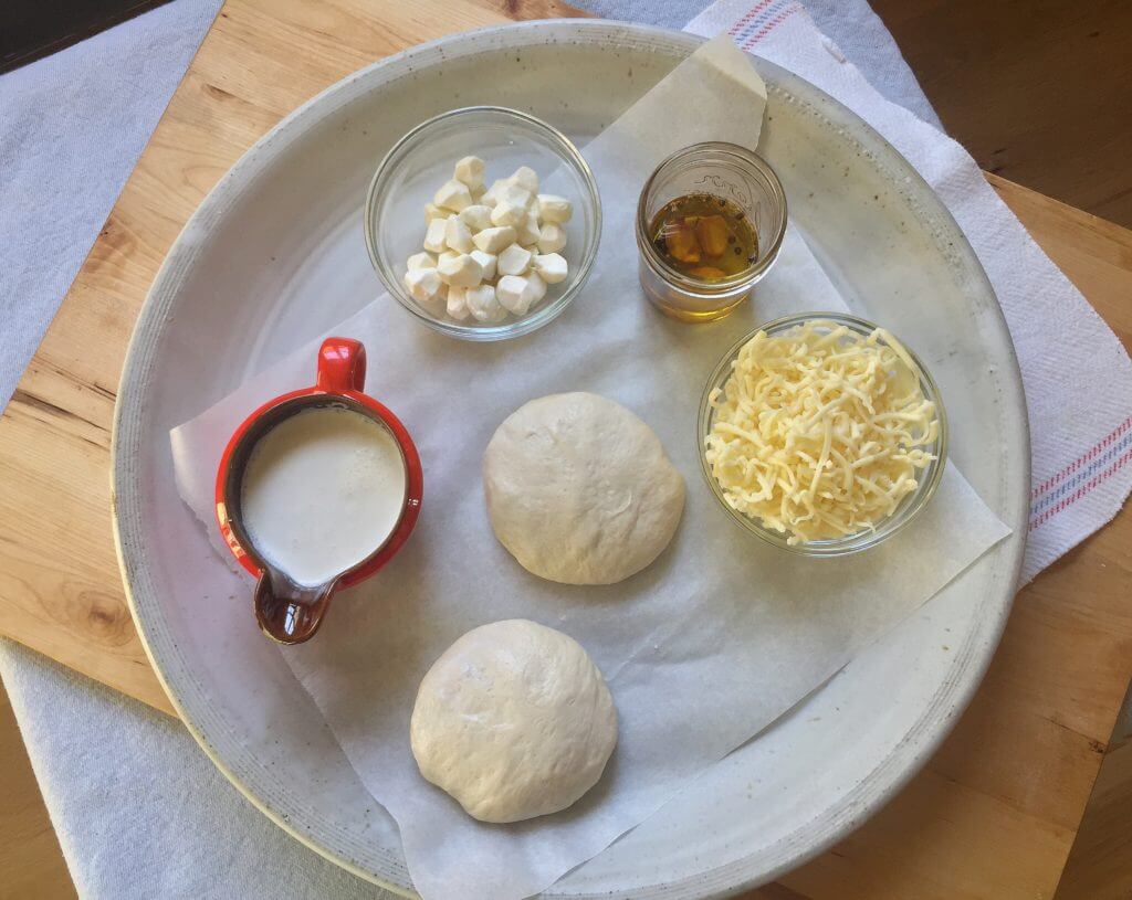 A tray with dough balls, shredded cheese, garlic oil, heavy cream, and small mozzarella cubes.