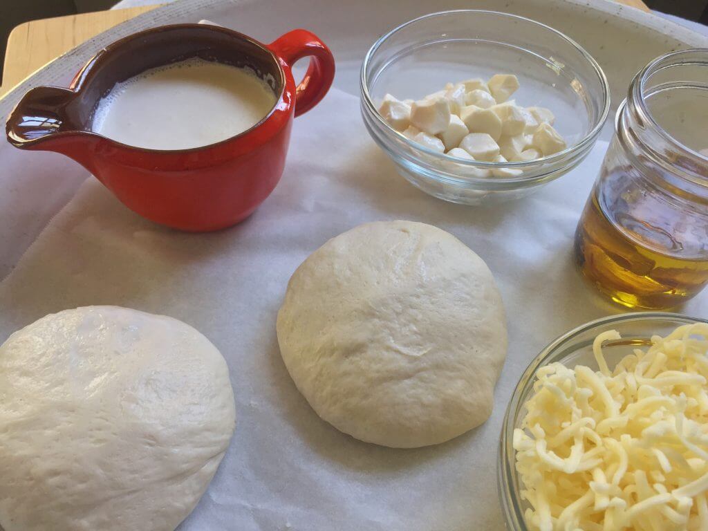 Three dough balls, shredded cheese, garlic oil, heavy cream in a red pitcher, and small mozzarella cubes on a tray.