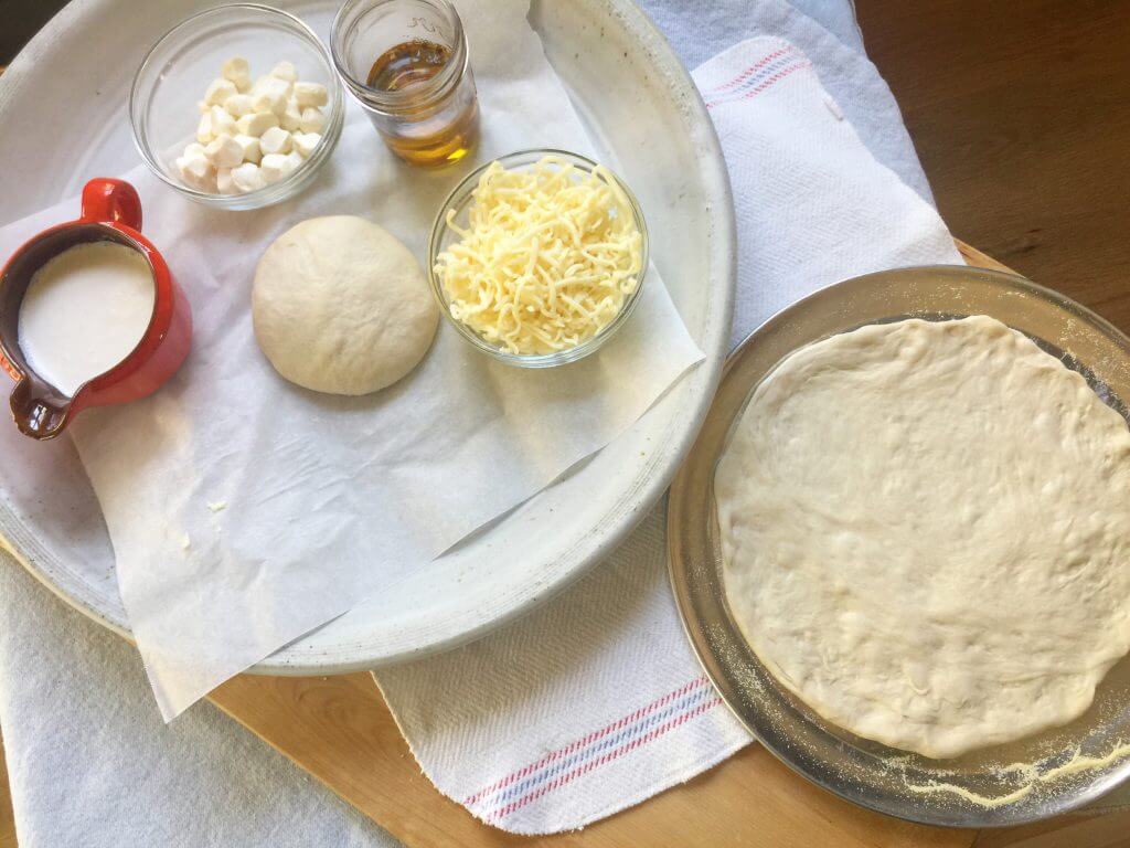 A tray with dough, shredded cheese, garlic oil, heavy cream, and small mozzarella cubes next to a rolled-out pizza dough.