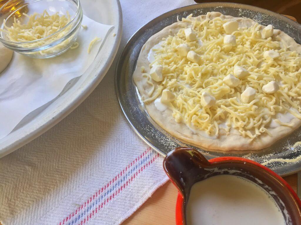 A pizza dough topped with shredded cheese, mozzarella cubes, and heavy cream next to a tray with more shredded cheese and a red pitcher of cream.