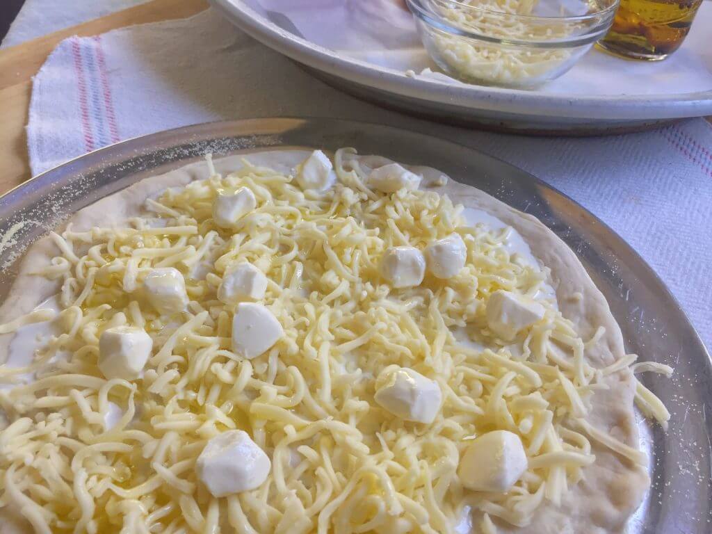 A close-up of a pizza dough topped with shredded cheese, mozzarella cubes, and heavy cream next to a tray with extra shredded cheese and a jar of garlic oil.