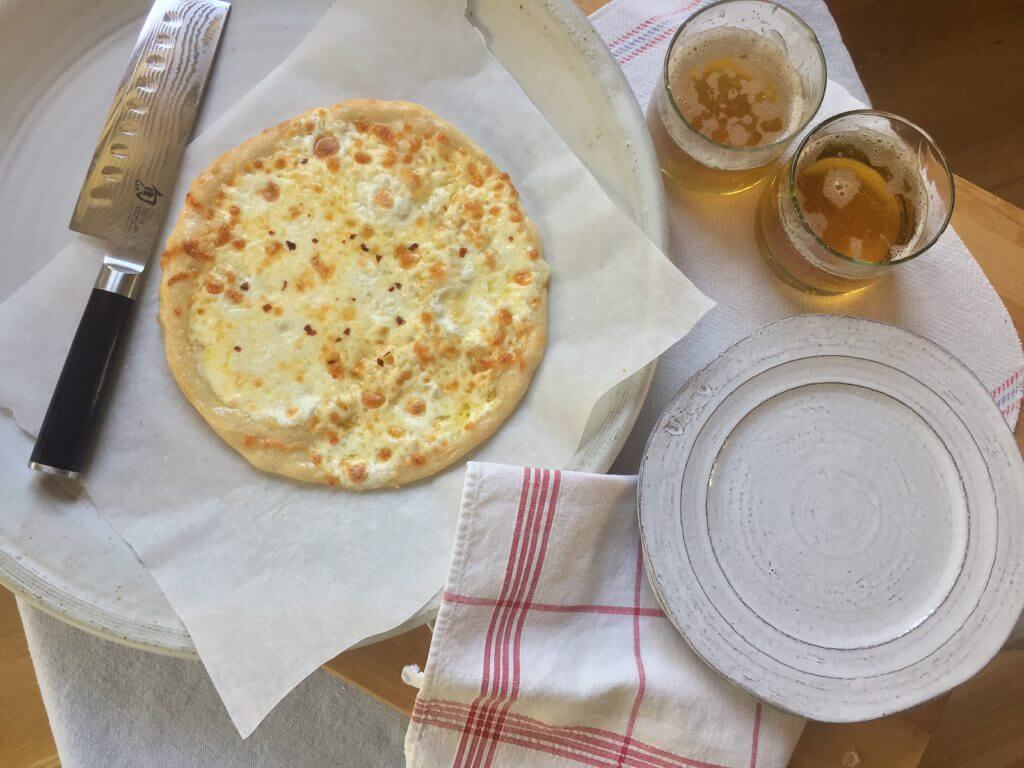 A cheese pizza on a tray with a knife, next to two glasses of beer and an empty plate on a table with a red and white napkin.