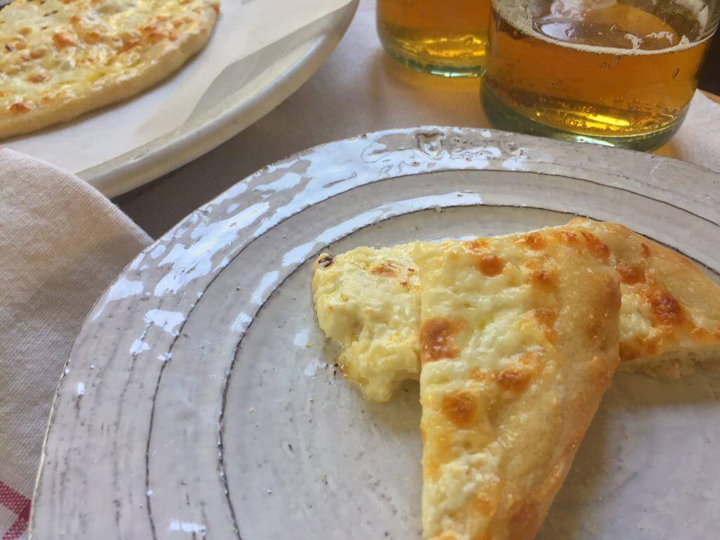 Two slices of cheese pizza on a plate with two glasses of beer in the background.