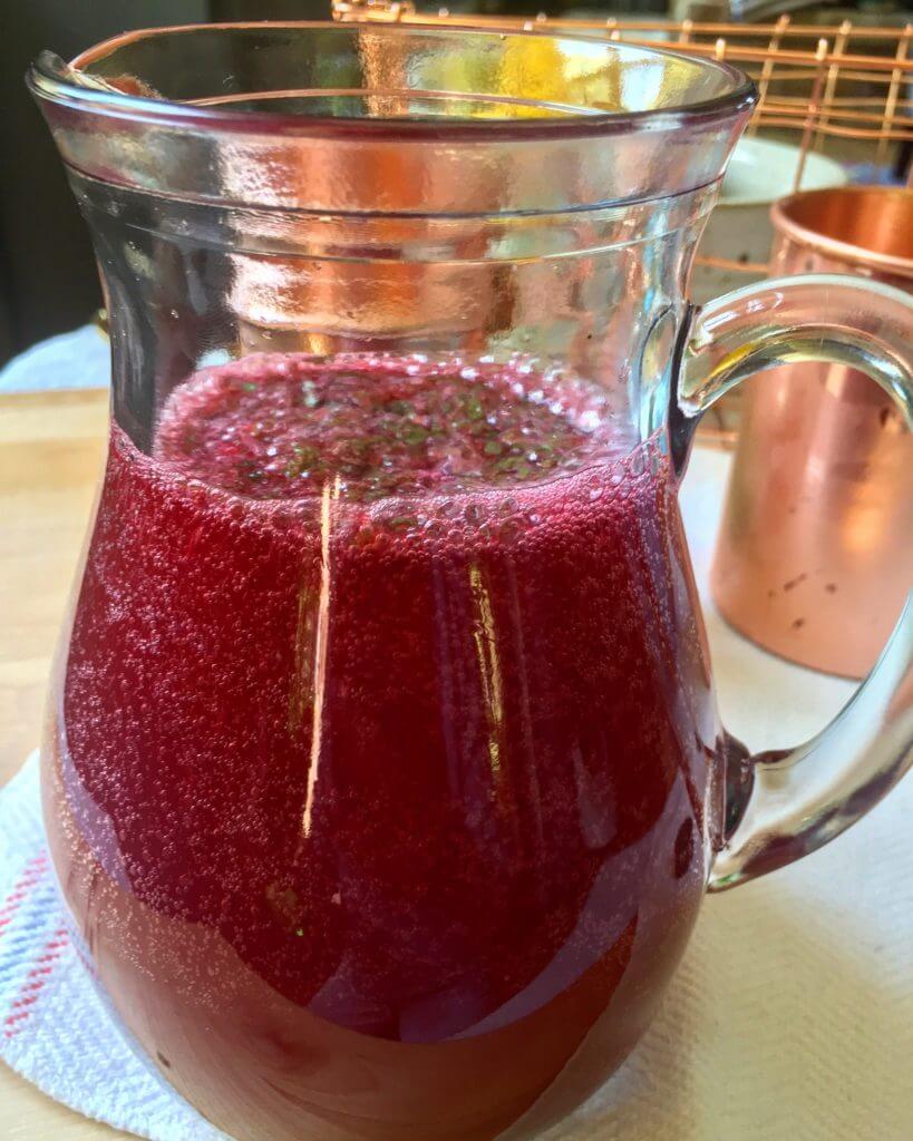 A glass pitcher filled with a bubbly red beverage topped with chopped mint, with copper mugs in the background.