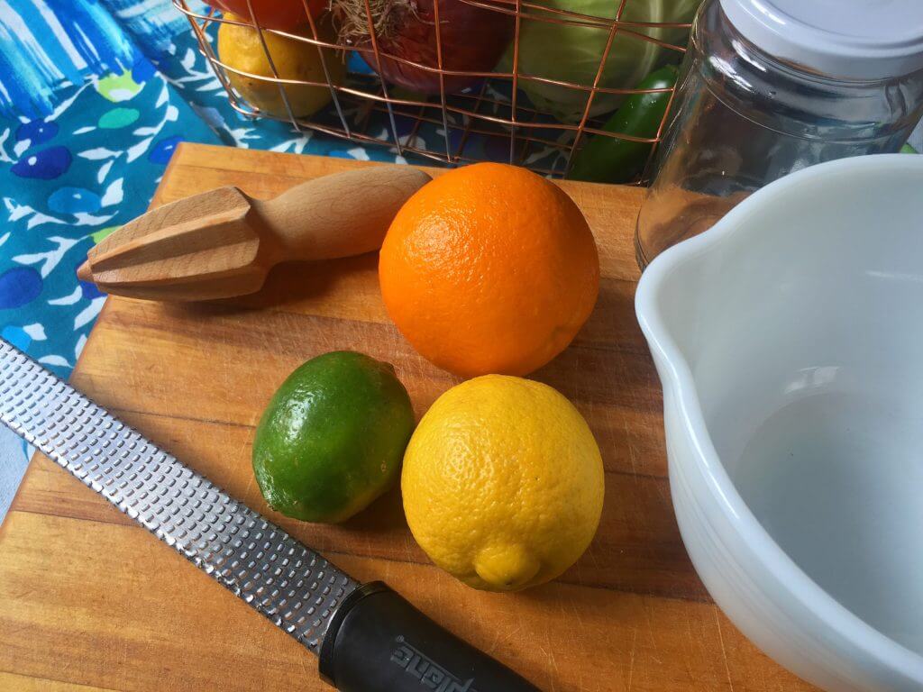 A wooden cutting board with an orange, a lemon, and a lime, a wooden citrus reamer, a grater, a white mixing bowl, and a wire basket containing various fruits and vegetables.