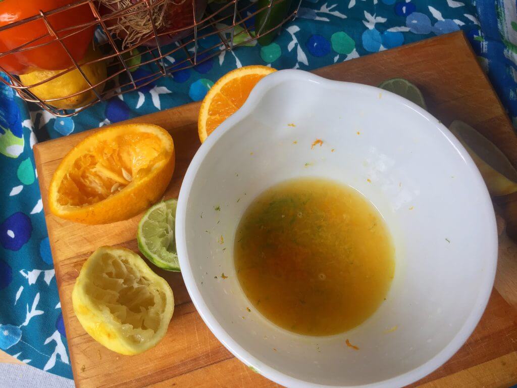 A white mixing bowl containing freshly squeezed citrus juice and zest, surrounded by squeezed orange, lemon, and lime halves on a wooden cutting board, with a wire basket of vegetables in the background.