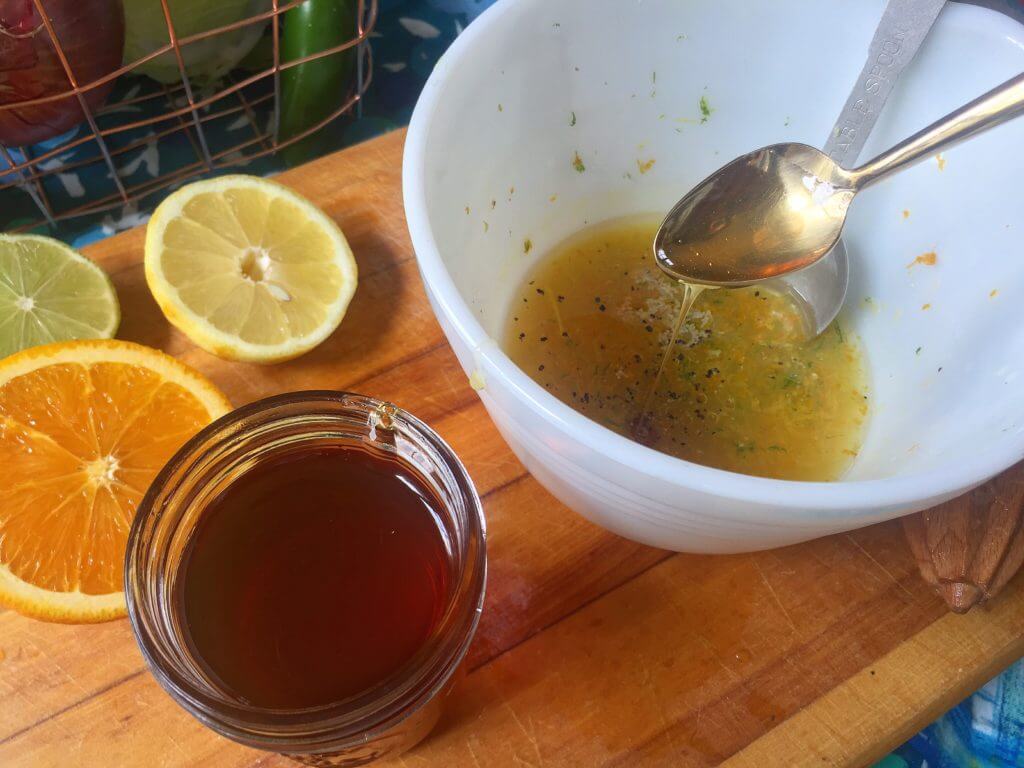 A white mixing bowl with citrus juice, zest, and seasonings being mixed with a spoon, next to halved citrus fruits (an orange, a lemon, and a lime) and a jar of honey on a wooden cutting board, with a wire basket of vegetables in the background.
