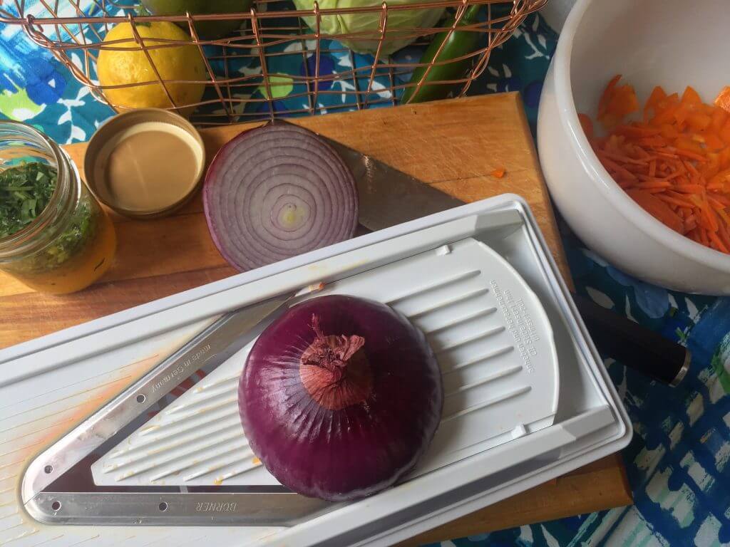 A red onion on a mandoline slicer on a wooden cutting board, next to a jar of chopped cilantro and citrus dressing, with a knife, a partially sliced onion, a lemon, and a bowl of grated carrots in the background, along with a wire basket of vegetables.