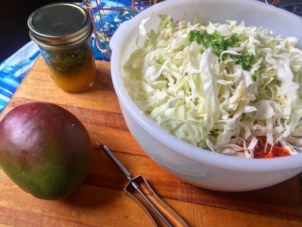 A white mixing bowl filled with shredded cabbage and grated carrots, topped with chopped cilantro and green onions, next to a jar of citrus dressing, a mango, and a vegetable peeler on a wooden cutting board, with a wire basket of vegetables in the background.