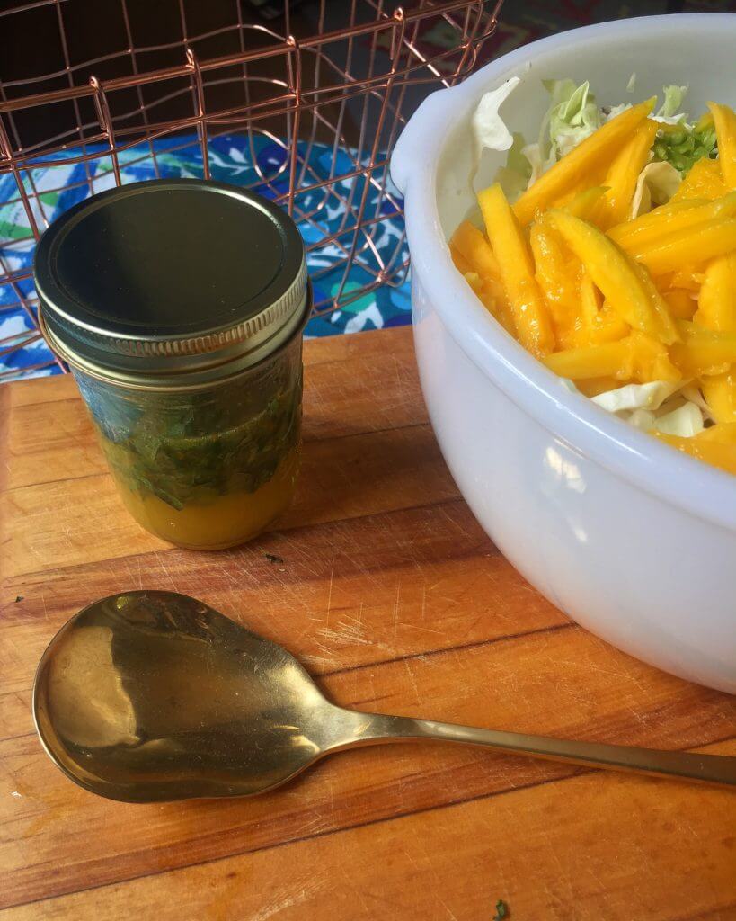 A jar of citrus dressing and a gold-colored spoon on a wooden cutting board, next to a white mixing bowl filled with shredded cabbage and sliced mango, with a wire basket of vegetables in the background.