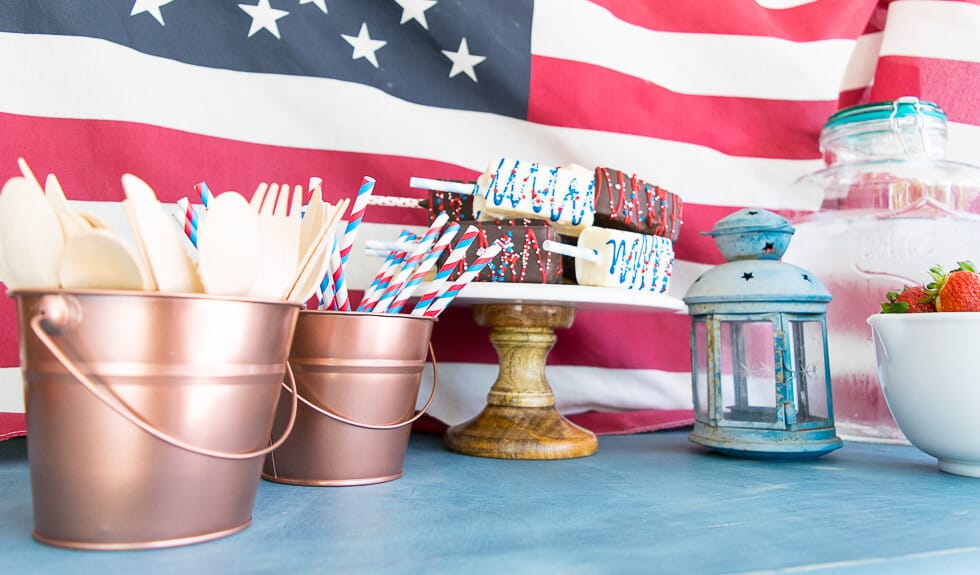 A patriotic-themed table setting featuring copper buckets filled with wooden utensils and striped straws, a wooden cake stand with decorated treats, a blue lantern, and a glass jar of pink lemonade, all set against an American flag backdrop. A bowl of strawberries is also visible.