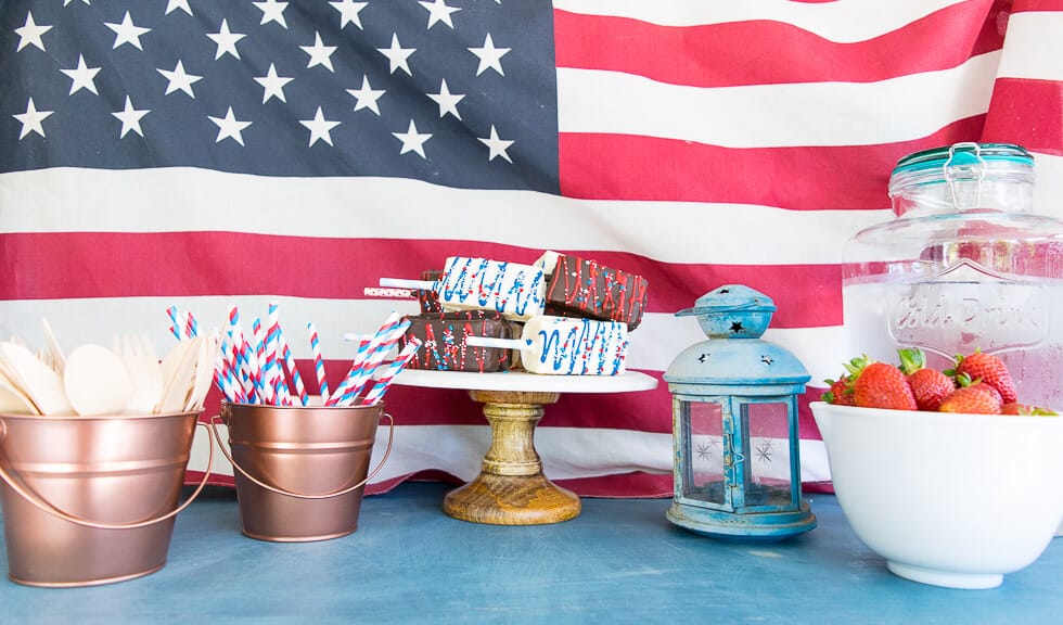 A festive Fourth of July display featuring a copper bucket filled with wooden cutlery and striped straws, a wooden stand holding decorated treats, a blue lantern, and a white bowl of strawberries. The backdrop is a large American flag.