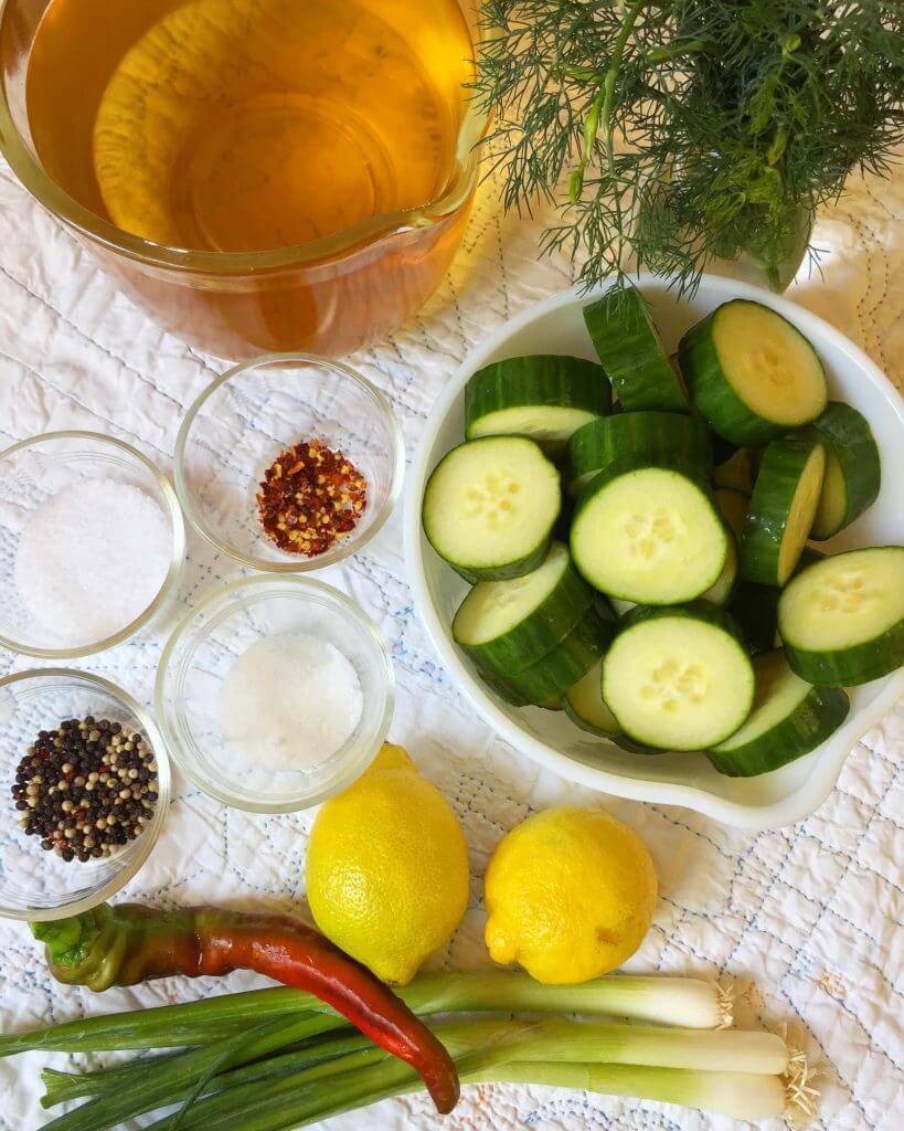 Ingredients for pickling including a bowl of cucumber slices, fresh dill, a pitcher of pickling brine, salt, peppercorns, red pepper flakes, lemons, green onions, and chili peppers arranged on a table.