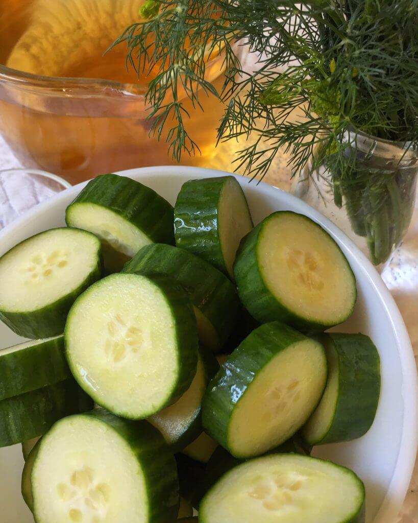 Bowl of cucumber slices next to a jar of fresh dill and a pitcher of pickling brine.