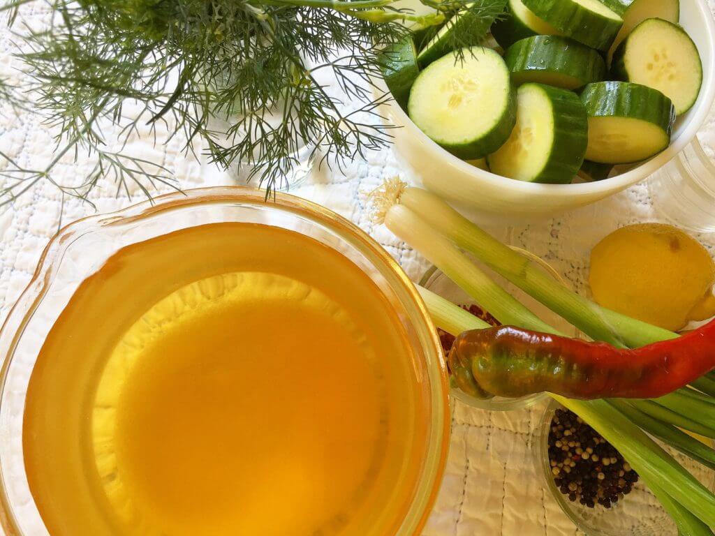 Ingredients for pickling including a bowl of cucumber slices, fresh dill, celery, lemon, a red chili pepper, and a jar of pickling brine.