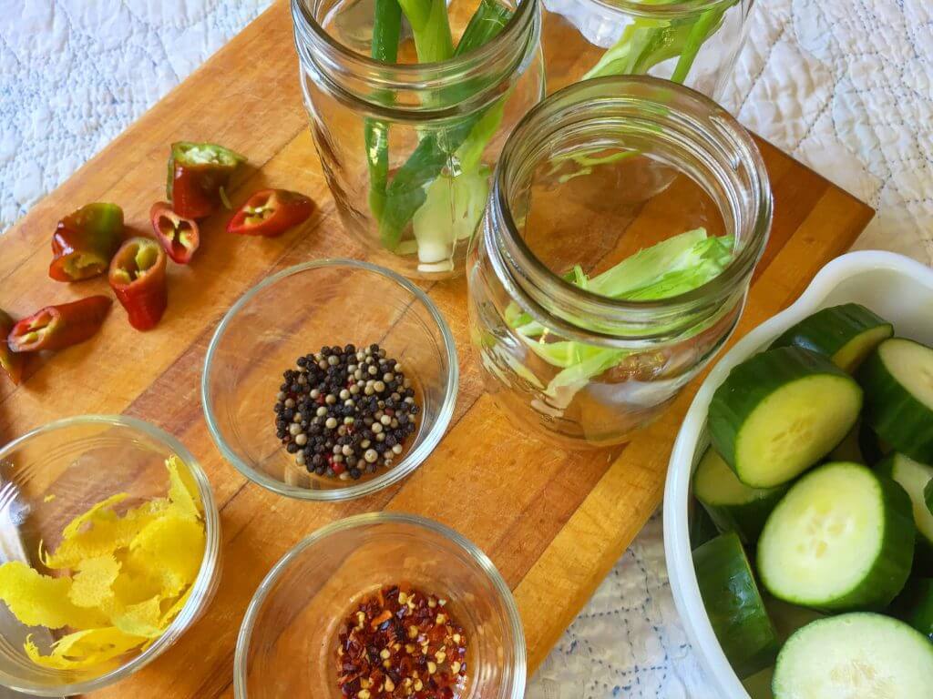 Preparation for pickling with jars containing celery, bowls of peppercorns, lemon peel, red pepper flakes, sliced chili peppers, and cucumber slices on a wooden surface.
