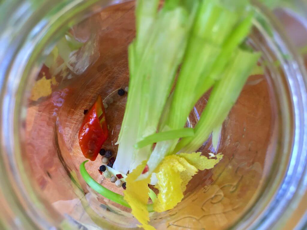 Top view into a jar with celery stalks, a red chili pepper, peppercorns, and lemon peel.