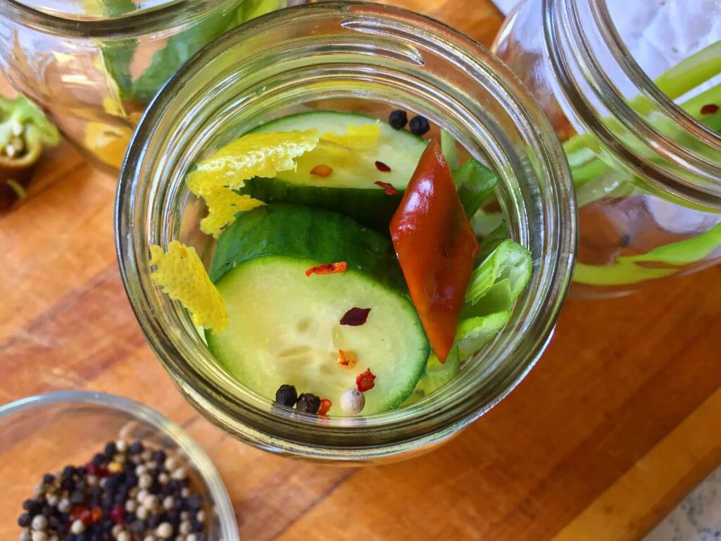 Top view of a jar containing cucumber slices, a red chili pepper, lemon peel, celery, and pickling spices.