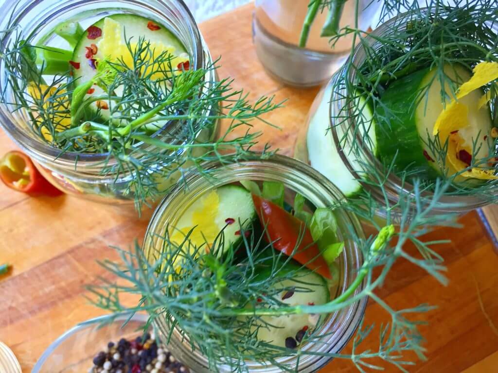 Top view of jars filled with cucumber slices, fresh dill, and pickling spices on a wooden surface.