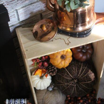A fall fireplace display with a "Halloween" banner, a crate of pumpkins, and a copper kettle with greenery.