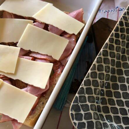 An open-faced sandwich in a baking dish topped with ham and slices of cheese, next to an empty patterned platter.