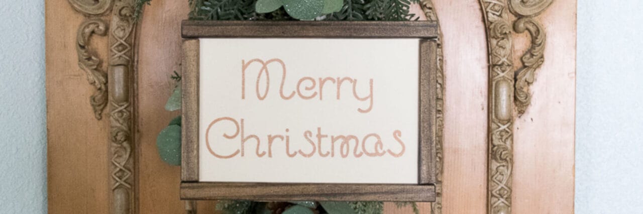 A sign with the handwritten phrase "Merry Christmas" is framed in wood and mounted on an ornate wooden background, with greenery peeking out from behind the frame.