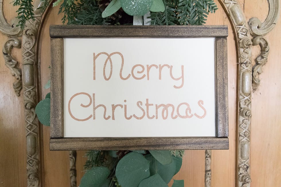 A "Merry Christmas" sign framed in wood is displayed against an ornate wooden background, with greenery accents behind the sign.