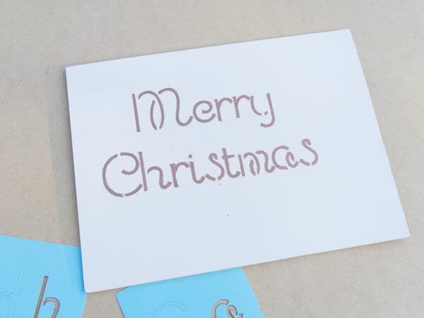The phrase "Merry Christmas" is handwritten in a casual, script-like font on a white surface, with blue stencils visible nearby.