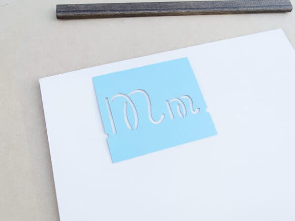 A blue stencil with the letters "M" and "m" cut out is placed on a white surface, with a wooden strip visible in the background.