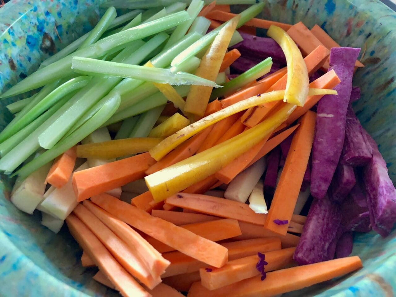 A bowl of sliced vegetables, including celery, carrots, yellow carrots, and purple sweet potatoes, ready for roasting.