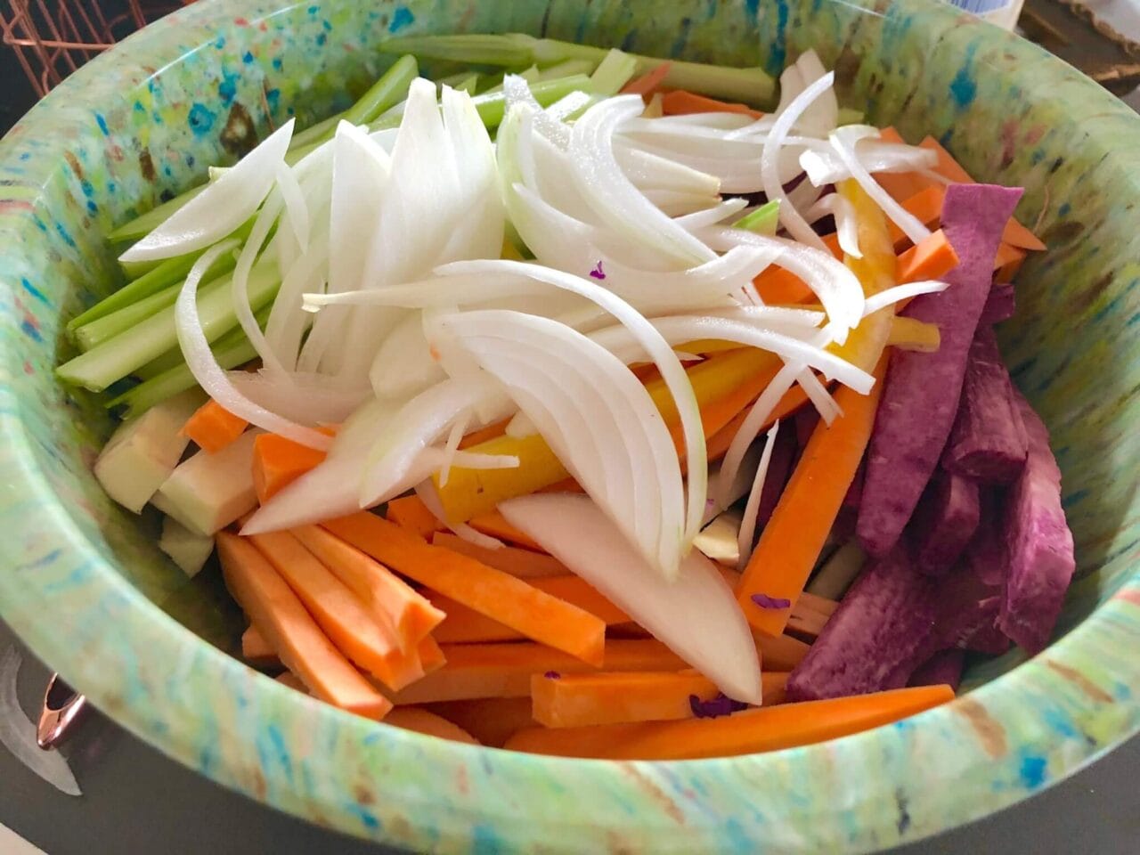 A bowl of sliced vegetables including celery, carrots, purple sweet potatoes, and onions, ready for roasting.