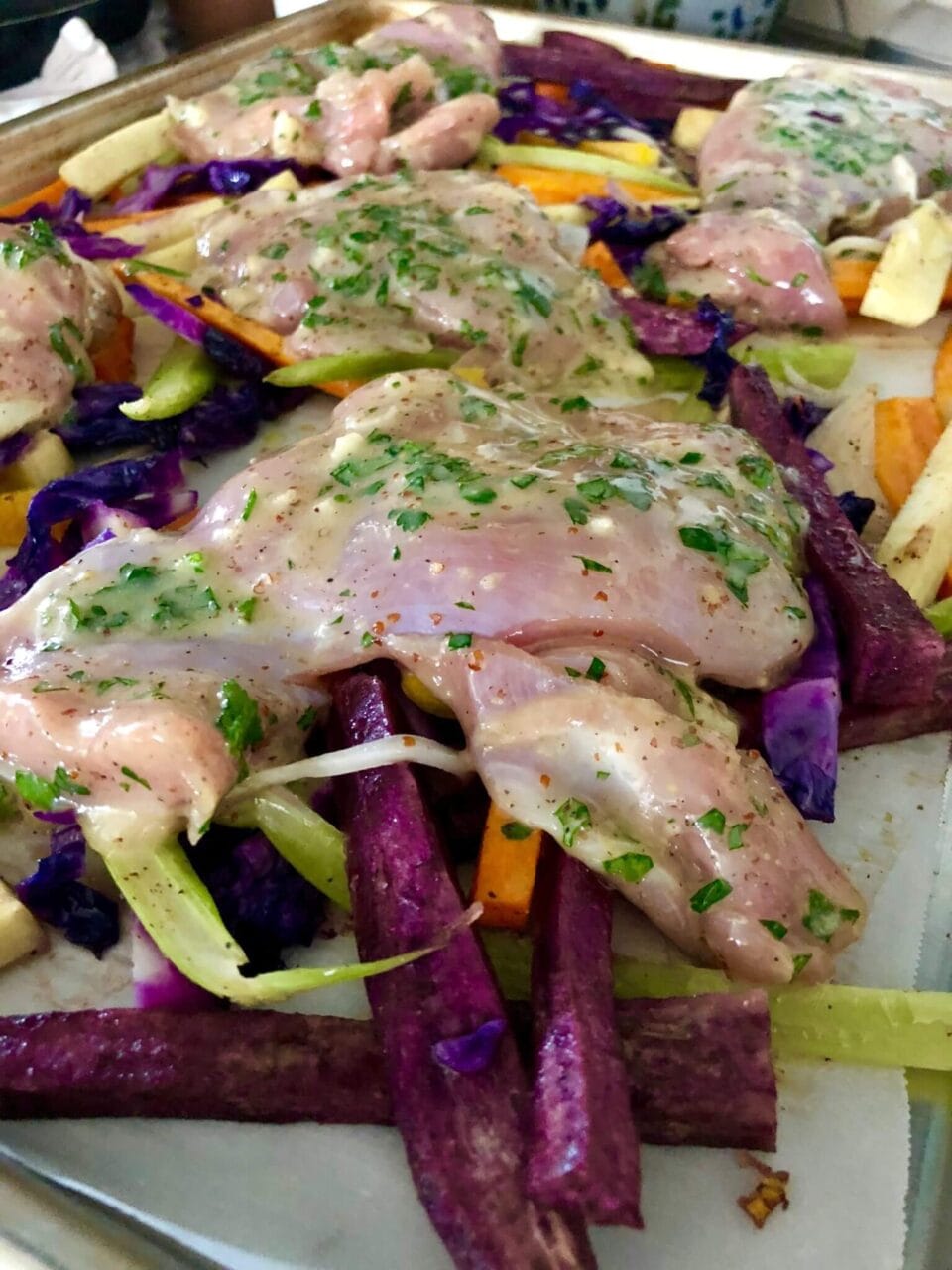 Raw honey mustard marinated chicken placed on a sheet pan with a variety of seasoned root vegetables, ready for roasting.