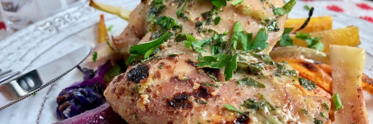 Close-up of roasted honey mustard chicken with root vegetables, garnished with fresh herbs, served on a white plate.