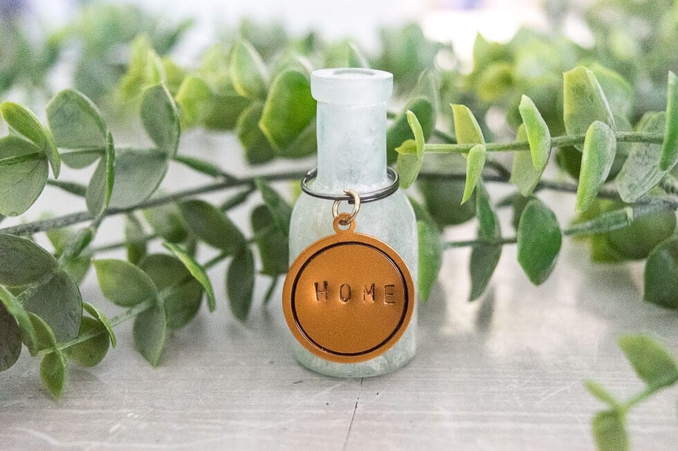 A small frosted glass bottle with a round metal tag stamped with the word "HOME," surrounded by green foliage. The tag is attached to the bottle with a keyring.