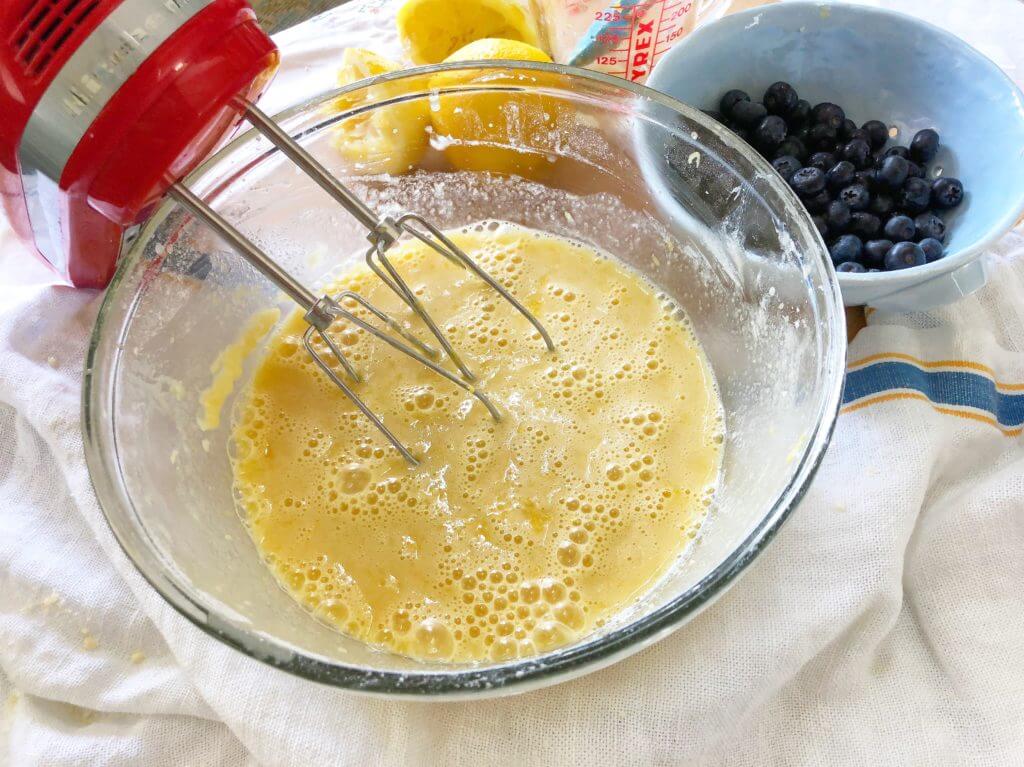 A glass bowl with a frothy lemon mixture being mixed by a hand mixer.