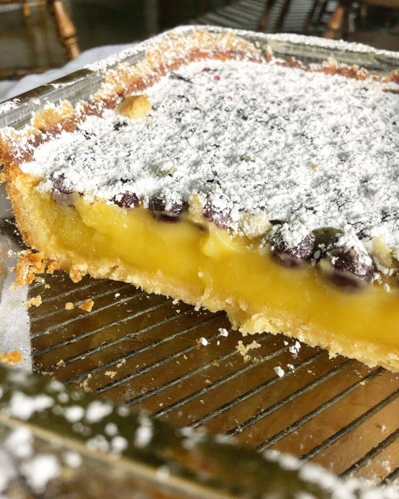 A close-up of a sliced lemon blueberry dessert with a powdered sugar topping in a glass baking dish, showing the creamy lemon filling and blueberries.
