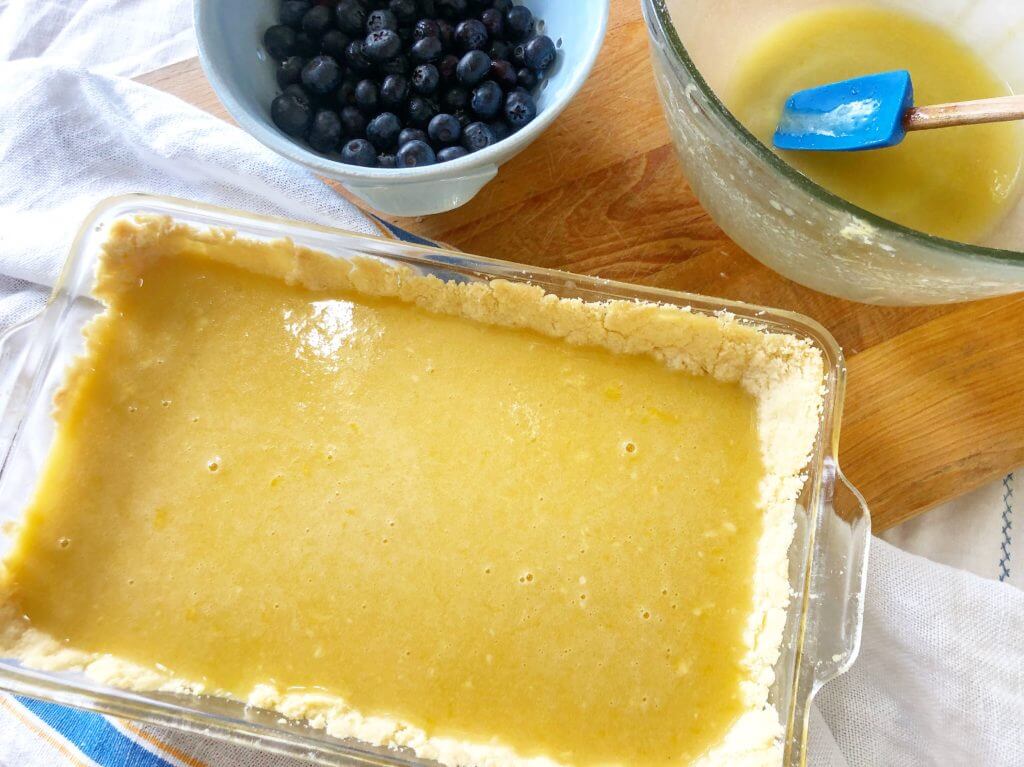 A glass baking dish with an unbaked lemon filling and crust, next to a bowl of blueberries and a mixing bowl with a spatula.