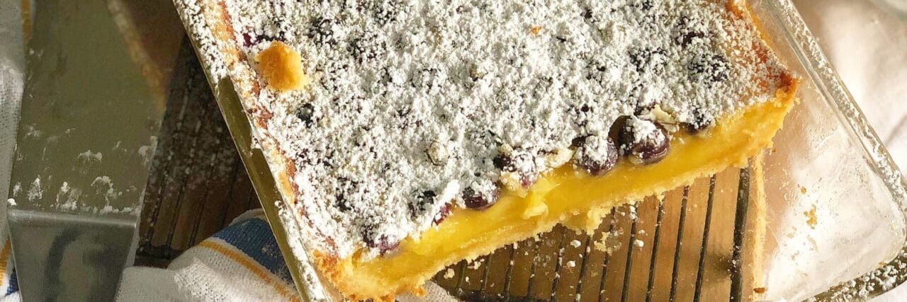 A close-up view of a baked lemon blueberry dessert topped with powdered sugar in a glass baking dish, partially sliced, on a cooling rack.