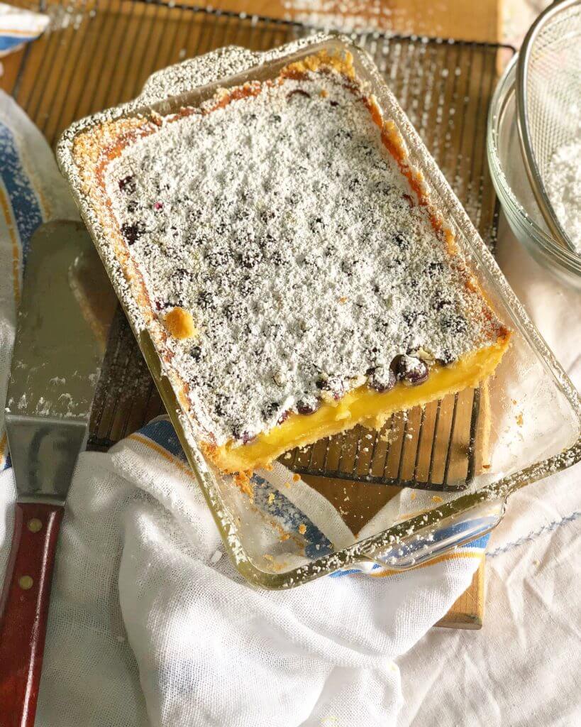 A glass baking dish with a baked lemon blueberry dessert topped with powdered sugar, partially sliced, resting on a cooling rack.
