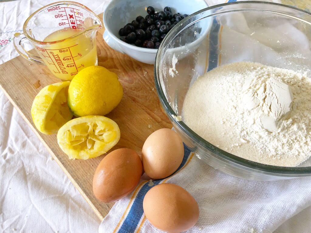 A wooden board with a glass bowl of flour, three eggs, lemon juice in a measuring cup, two squeezed lemons, and a bowl of blueberries.