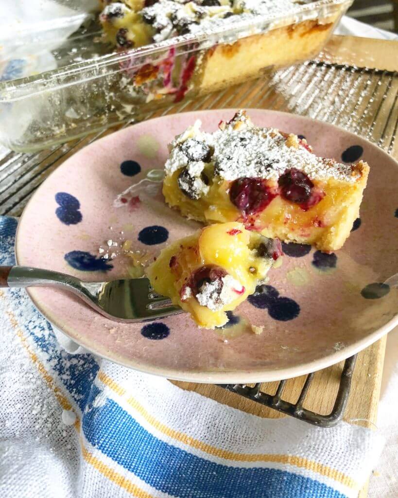 A slice of blueberry lemon dessert dusted with powdered sugar on a pink plate with blue polka dots, with a fork holding a bite and a glass baking dish in the background.