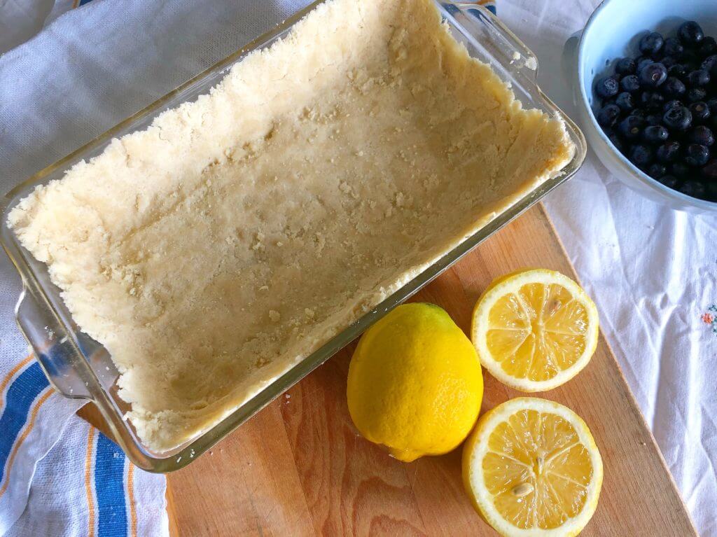 A glass baking dish with an unbaked crust, placed on a wooden board next to halved lemons and a bowl of blueberries.