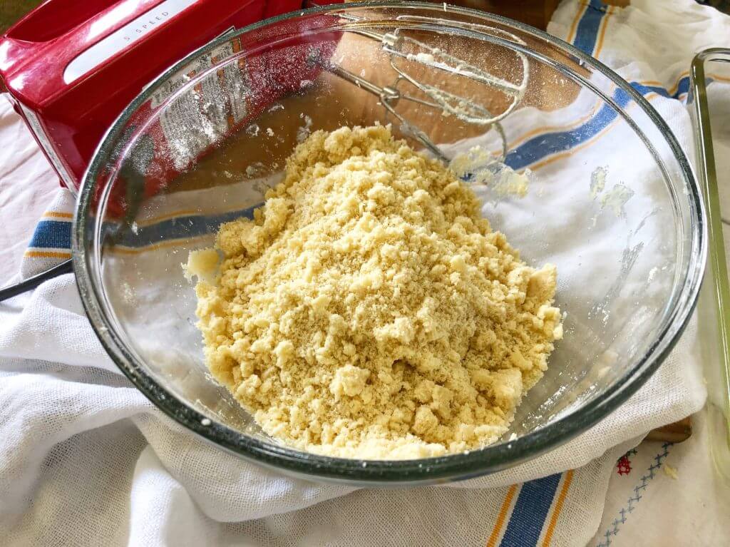 A glass bowl containing crumbly dough mixture, with an electric mixer and a red mixer base visible in the background.