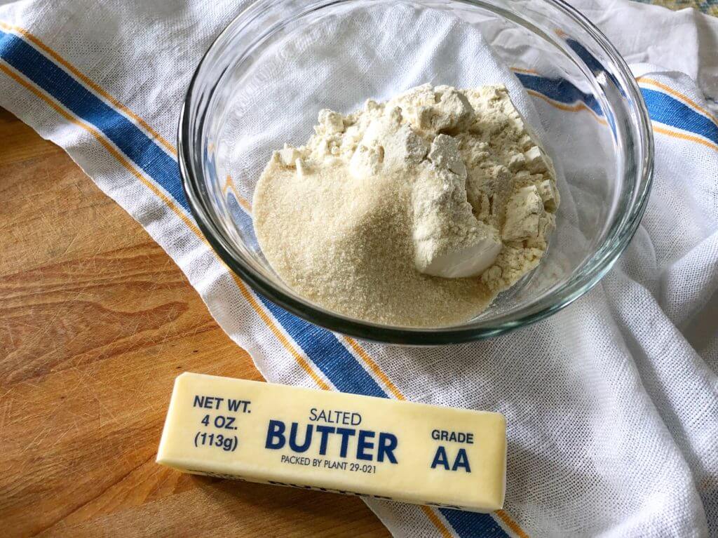 A glass bowl containing flour and sugar, next to a stick of salted butter, placed on a wooden board with a striped cloth underneath.